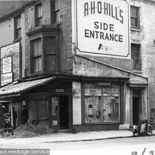 Street corner with a large sign on the wall showing directions to the R.H.O. Hill's side entrance, using an illustrated hand pointing to the right.