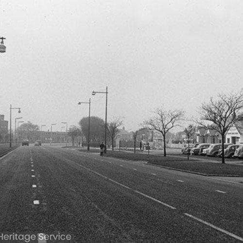 Road lined by street lights with buildings and parked cars on the right.