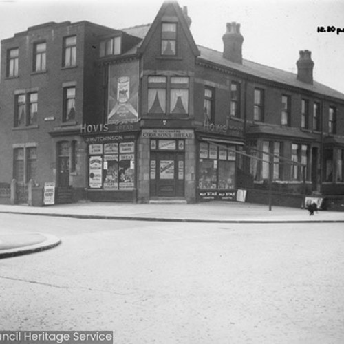 Street corner and the shop front of J.Hutchinson with advertisements for Hovis and Cookson's Bread.
