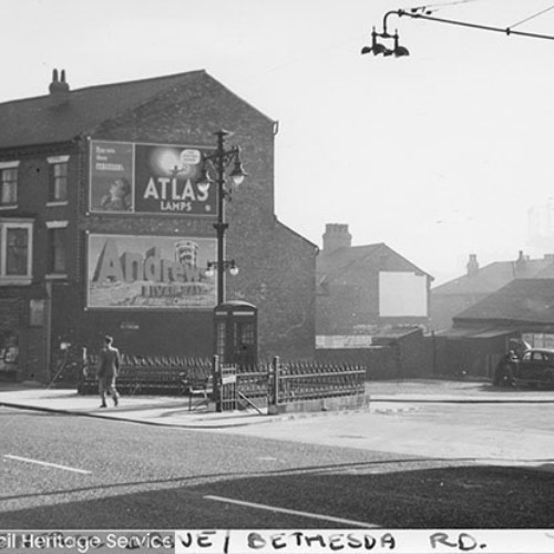 Street corner, with shop fronts and the side wall of the end building covered in poster advertisements. A public telephone box is also on the corner.