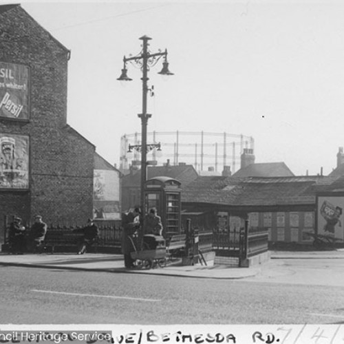 Street corner, with the end building covered in poster advertisements for Persil and Andrews. There is also a square area where people are sat on the benches and also a Public Telephone Box.