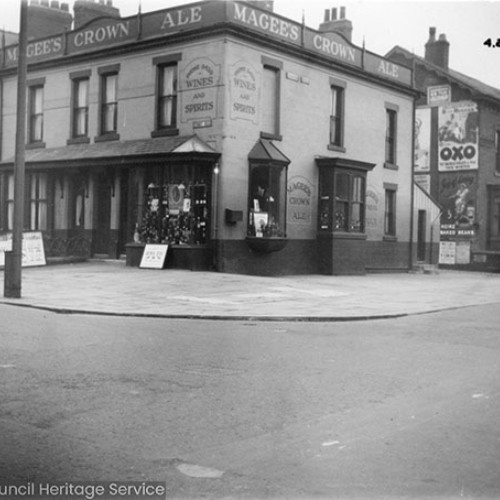 Street corner, with the off licence on the corner advertising Magee's Crown Ale. On the side wall of the buildings next to the off licence are a number of large advertising posters for brands such as OXO.