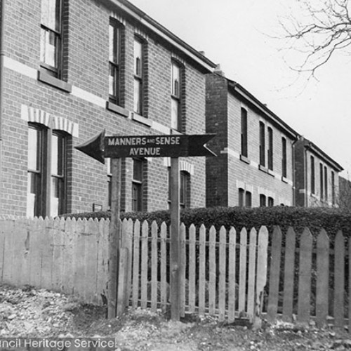 Street sign with an arrow pointing towards Manners and Sense Avenue. A fence and a row of houses are behind the sign.