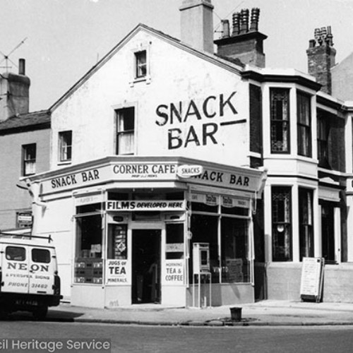 Street corner, with the Corner Cafe/Snack Bar on the corner which is advertising tea, coffee, snacks and film developing.