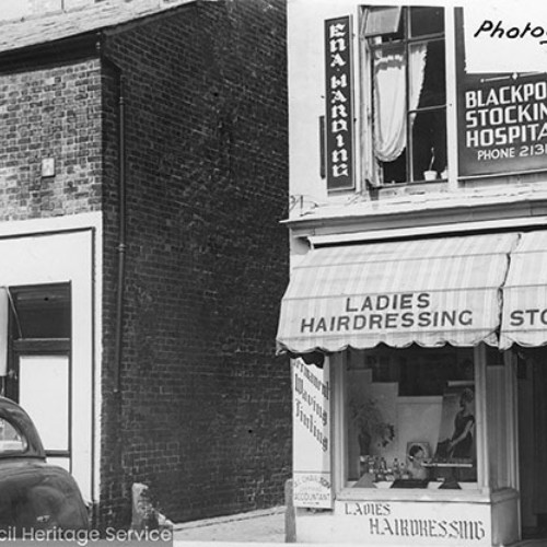 Two buildings separated by an alleyway. The building to the right is advertised as both a Ladies Hairdressing salon and the Blackpool Stocking Hospital.