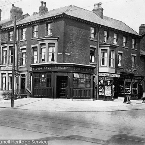 Street corner with a Lloyds Bank on the corner and a number of other shop fronts on either side of the bank.