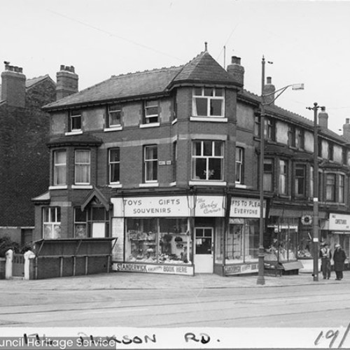 Street corner, with a shop on the corner advertising toys, gifts and souvenirs. Other shop fronts are to the right.