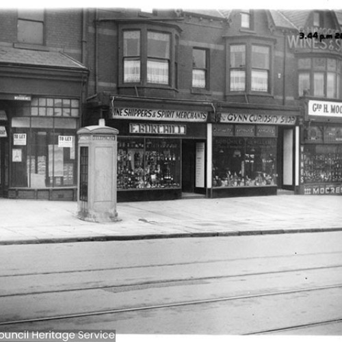 Shop fronts from left to right, there is a shop to let, F.Burchill Wine Shippers and Spirit Merchants, Gynn Curiosity Shop and G.H. Moore's Wine and Spirits.