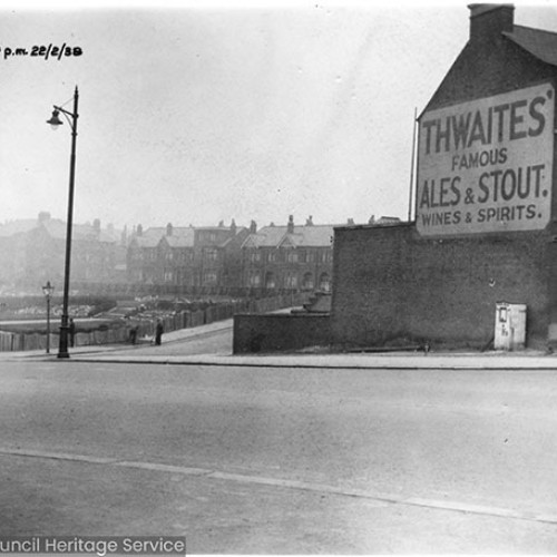 Side wall of a public house, which has a large advertisement on for Thwaites Famous Ales and Stout, Wines and Spirits. To the left of the public house is a large park area, with houses that run along the back.