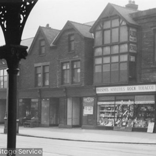 Row of shop fronts, with the shop on the street corner advertising presents, novelties, rock and tobacco.