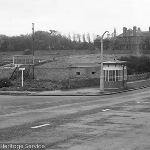 Field at the side of the road and a military Pill Box. There is a sheltered bus stop on the roadside and houses in the distance.