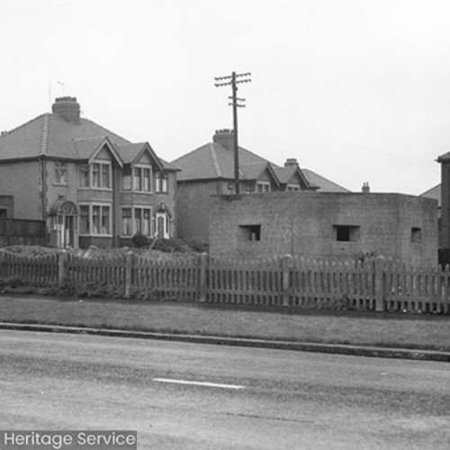 A Military Pill Box on a fenced off patch of land, with a number of houses visible on the road behind the Pill Box.