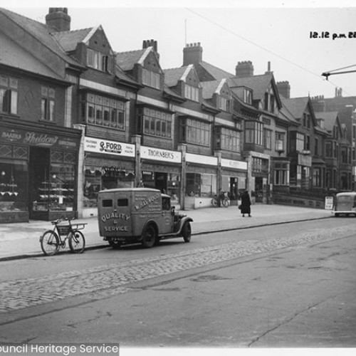 Shop fronts including Sheldon's, S. Ford and Thornber's. There is a W. Haywood butchers delivery van and bicycle parked on the road in front of the shops.