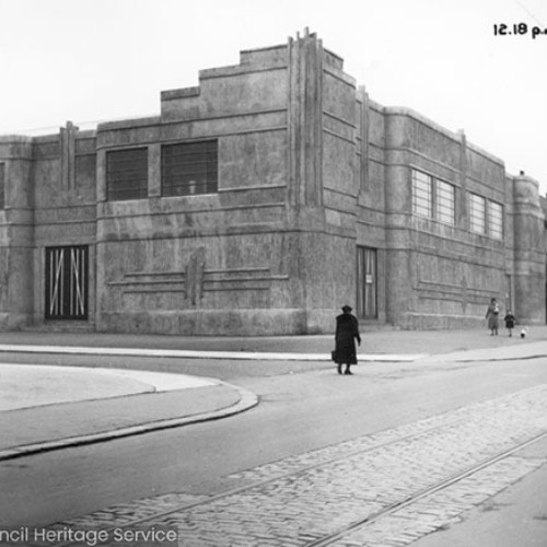 People walking past a large building on the corner of the road.