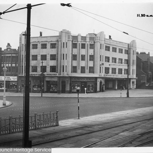 Street corner, with a large building on the corner which is occupied by shops on the ground floor including a chemist. There is a road in front of the building and then there are tram lines with overhead wires in front of the road.