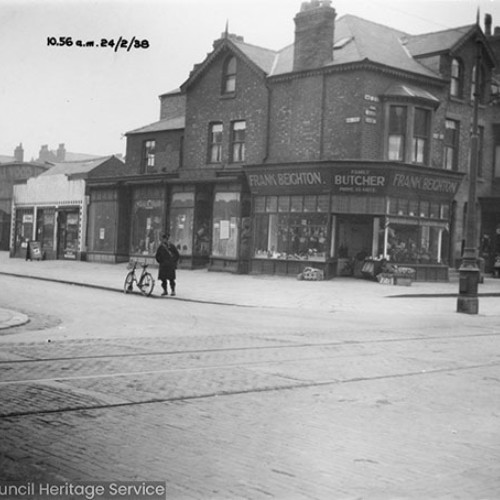 Street corner with Frank Beighton Family Butcher on the corner and a man stood next to a parked bicycle on the road.