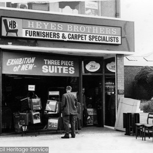 Shop front of Heyes Brothers Furnishers and Carpet Specialists, with four chairs and some other furniture on the forecourt. A man is looking through the shop window, as another man is exiting the shop.