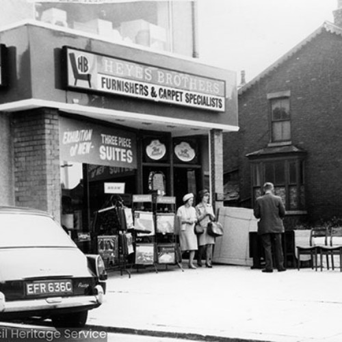 Shop front of Heyes Brothers Furnishers and Carpet Specialists with a man stood by four chairs and other furniture on the forecourt and two ladies exiting the shop.