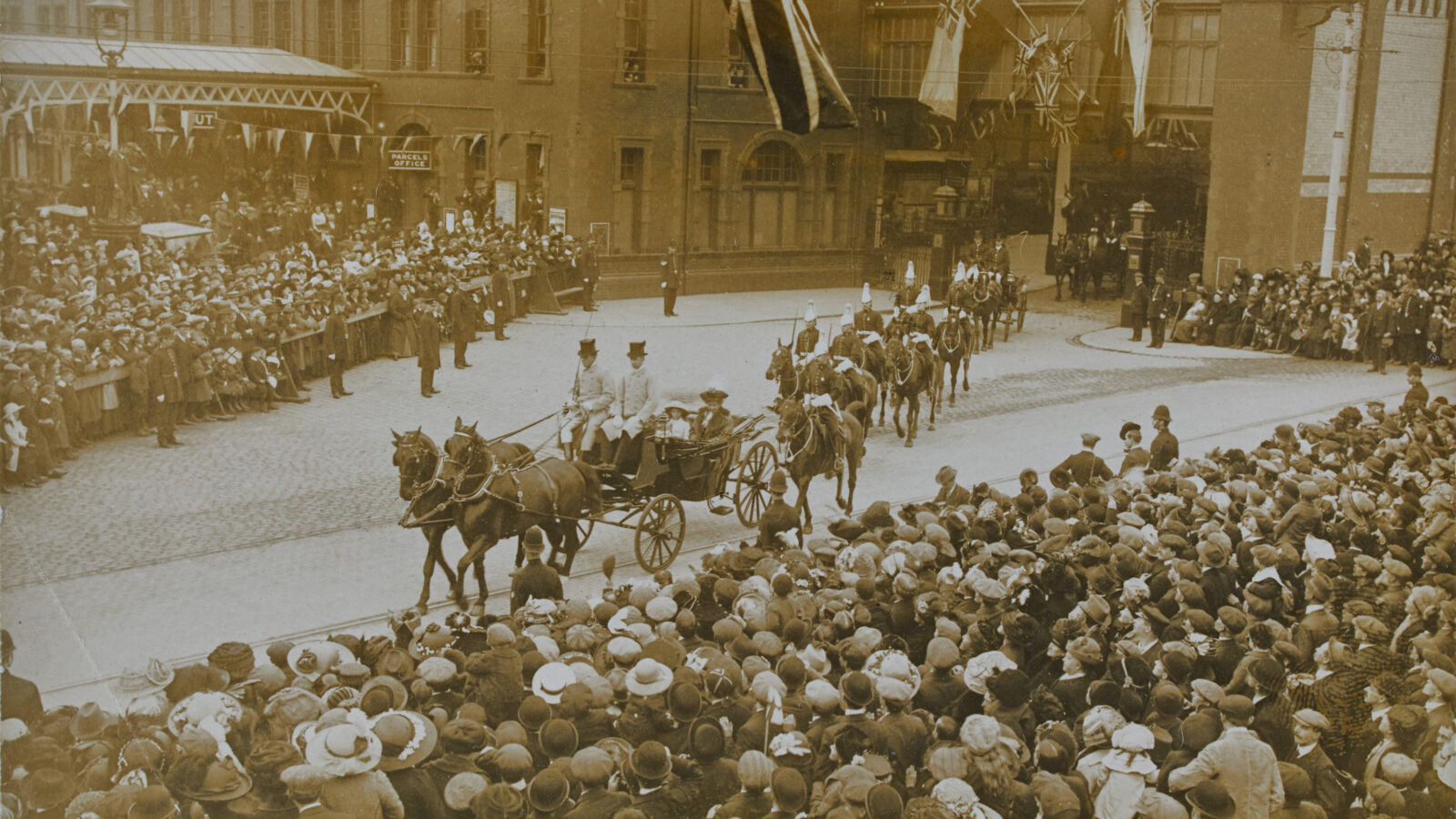 Black and white photograph of horse and carriage leaving the train station. Crowds line the streets.