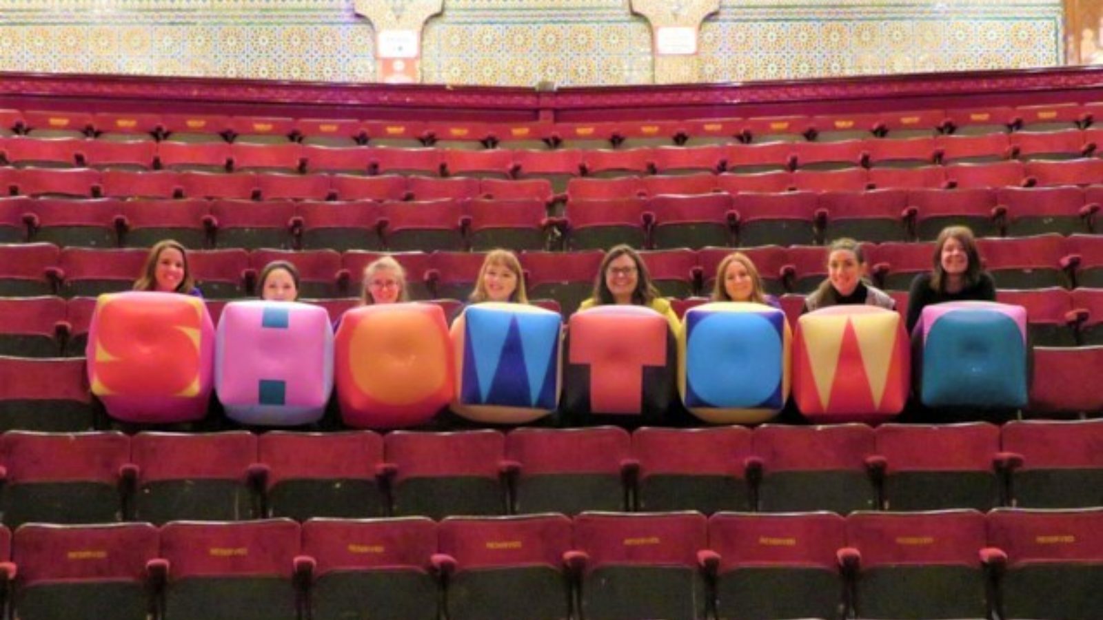 Showtown team, surrounded by theatre chairs, holding inflatable letters spelling Showtown.