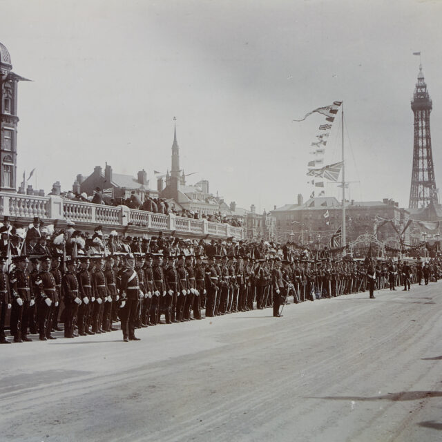 Black and white photograph showing the parade lined with crowds and soldiers. Blackpool Tower can be seen in the background.