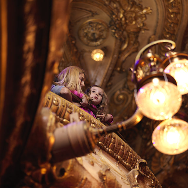 Image of a woman and a young girl leaning over the balcony of a theatre.  Their faces are illuminated by 3 bright lights