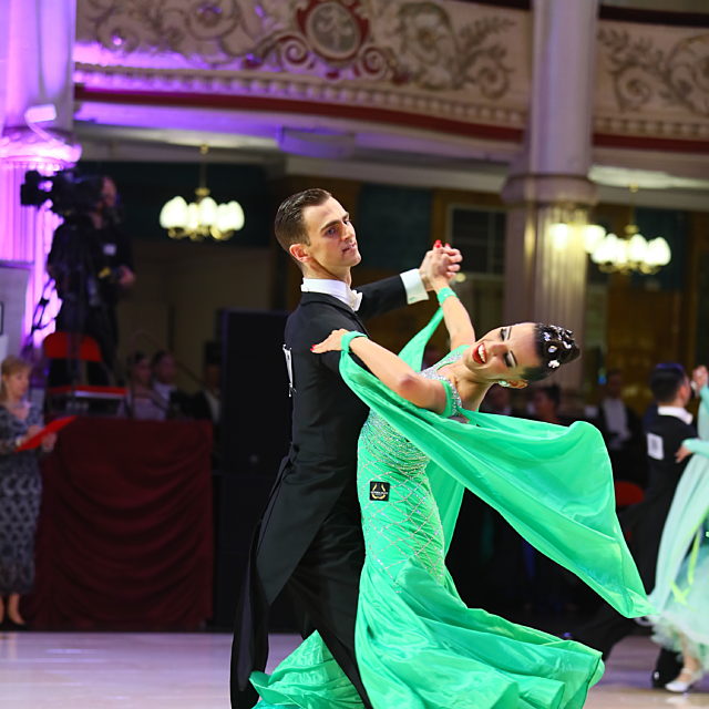 A pair of ballroom dancers dancing. She wears a green floating dress and he wears a suit.