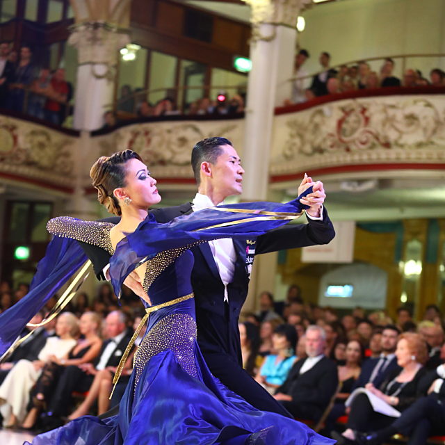 A pair of ballroom dancers.  She wears a long purple gown and he wears a suit.