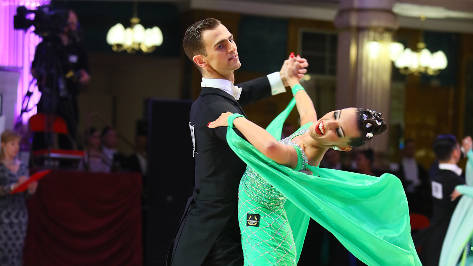 A pair of ballroom dancers dancing. She wears a green floating dress and he wears a suit.