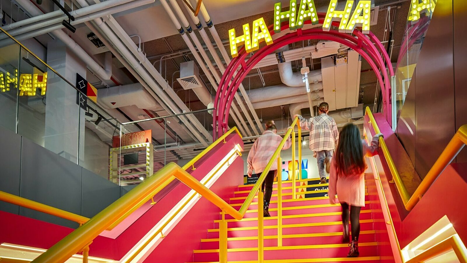 Three girls walking up a bright pink staircase at the entrance of the Showtown Blackpool museum.  The word Ha, ha, ha are spelled out in yellow lights.