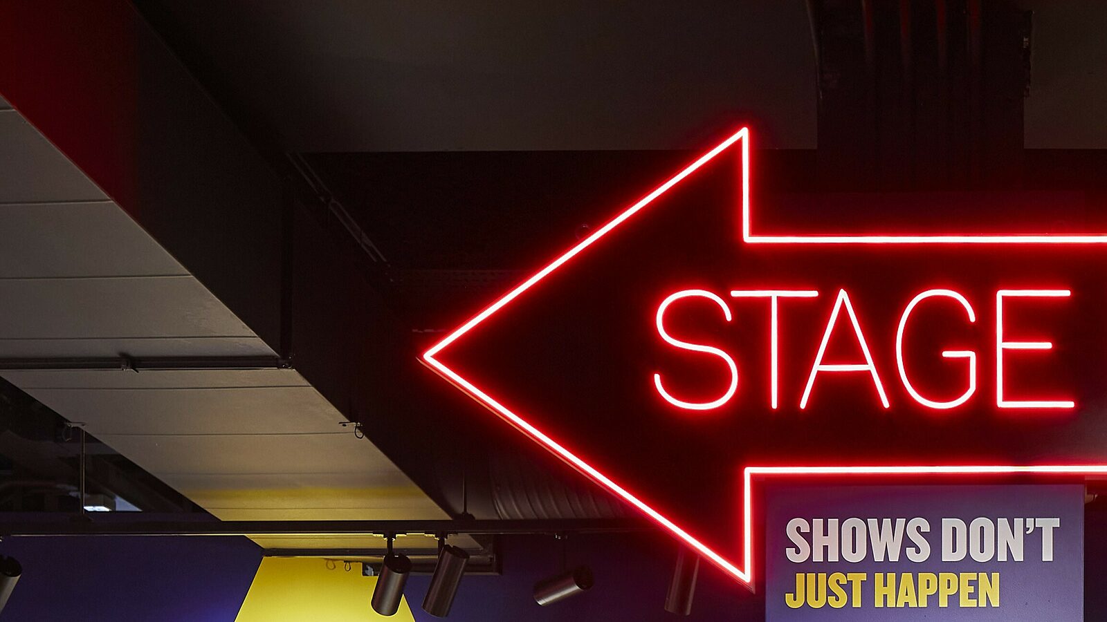A neon arrow shaped sign with the word stage inside. Text below reads 'Shows Don't Just Happen'