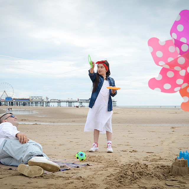A lady and girl on the beach relaxing and playing with toys. A colourful wind mill sits in front