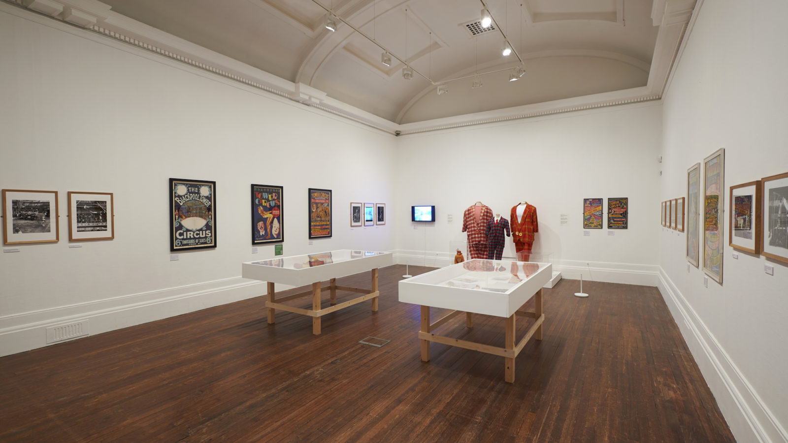 A white wall gallery, with images hung around the room and three suits in the background.