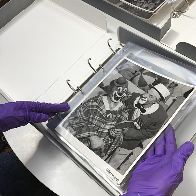 A pair of hands wearing purple gloves, holding a old photo of two clowns.