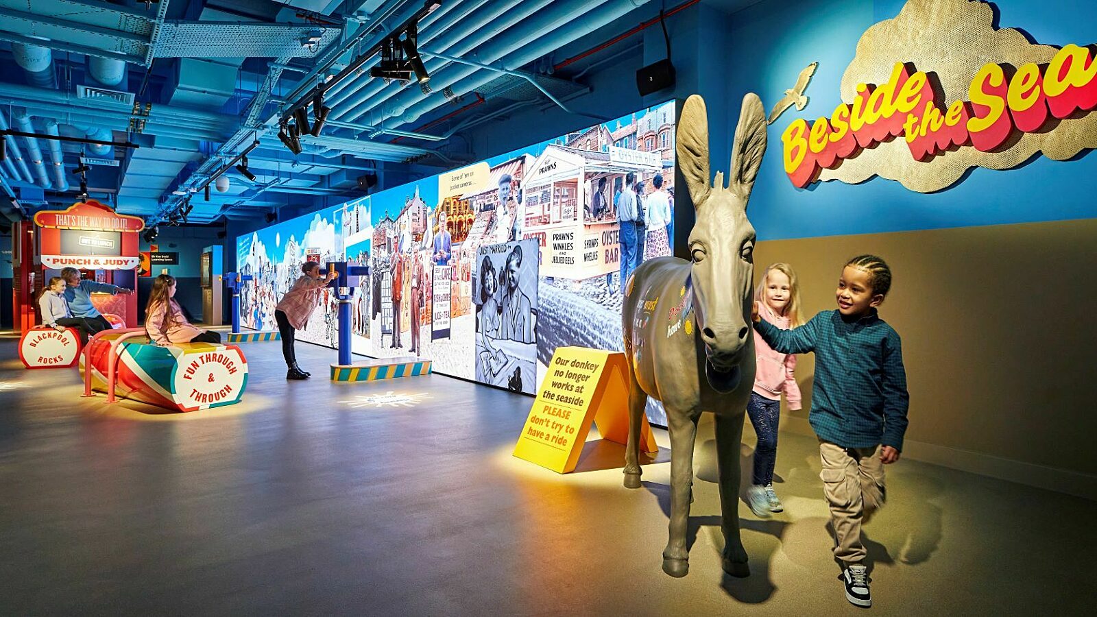 Seaside Gallery.  Children play around a model donkey, child looks through telescope at large interactive screen. Punch and Judy in background.