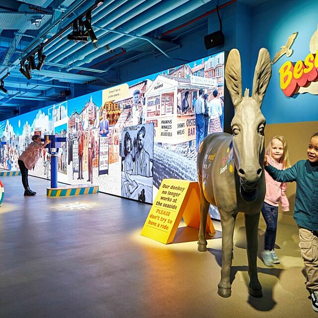 Seaside Gallery.  Children play around a model donkey, child looks through telescope at large interactive screen. Punch and Judy in background.